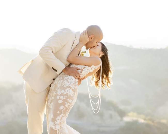 A couple kisses while standing on a hillside, dressed in wedding attire, with a scenic landscape in the background.