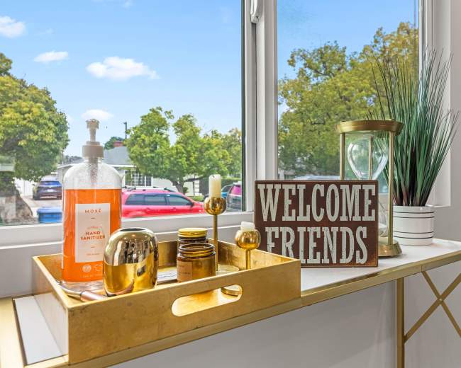 A decorative table features a welcome sign, various bottles, a candle holder, and a plant, all set near a window with a view of trees and parked cars outside.