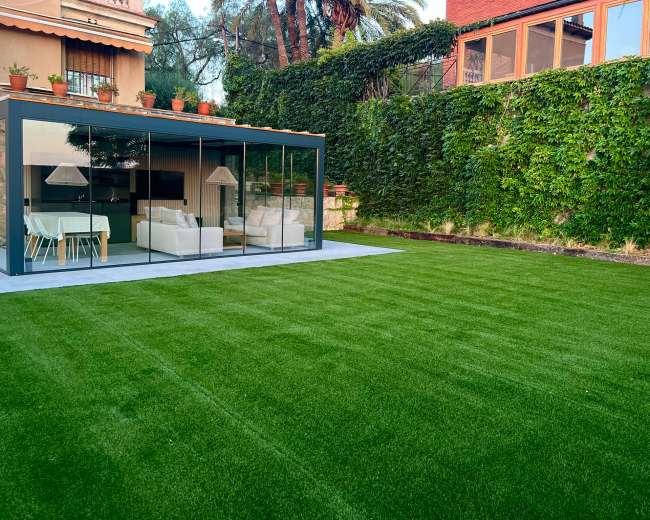 The image shows a modern glass-enclosed living space overlooking a green lawn with a wall of greenery in the background.