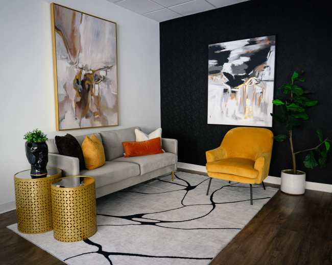 The image shows a modern seating area featuring a gray sofa with decorative pillows, a bright yellow chair, two gold patterned side tables, and two abstract paintings on the walls, all placed on a patterned rug.