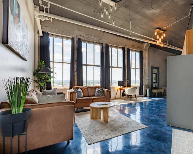The image shows a modern, open living space with large windows, brown sofas, a circular wooden coffee table, and polished blue flooring.