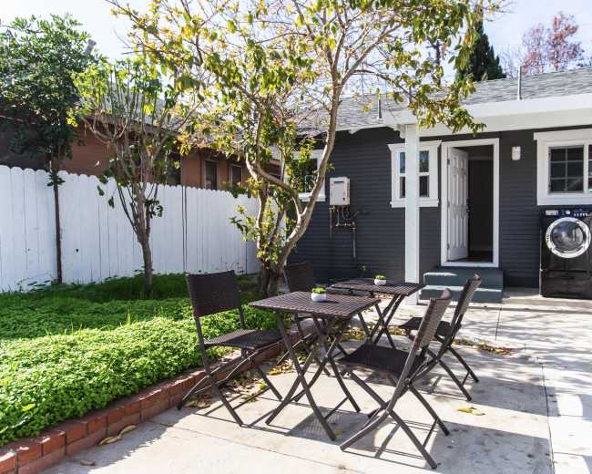 A small outdoor patio features a black metal table and chairs surrounded by greenery, with a washing machine and dryer visible nearby under a gray house exterior.