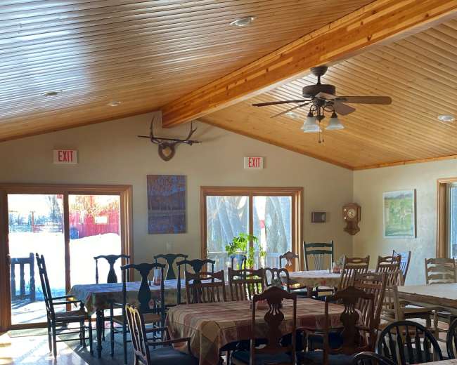 A spacious dining area with wooden tables and chairs, a high wooden ceiling, and large windows letting in natural light.