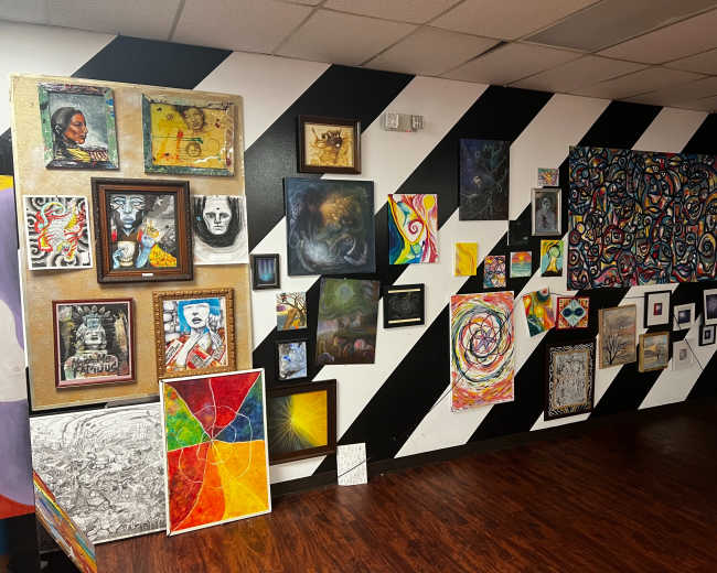 A wall with a variety of framed artwork and paintings in different styles and colors arranged against a striped black and white background.