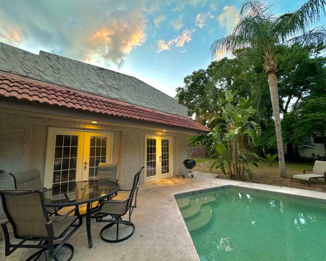 A patio area with a table and chairs overlooks a swimming pool surrounded by palm trees and tropical plants near a house at dusk.
