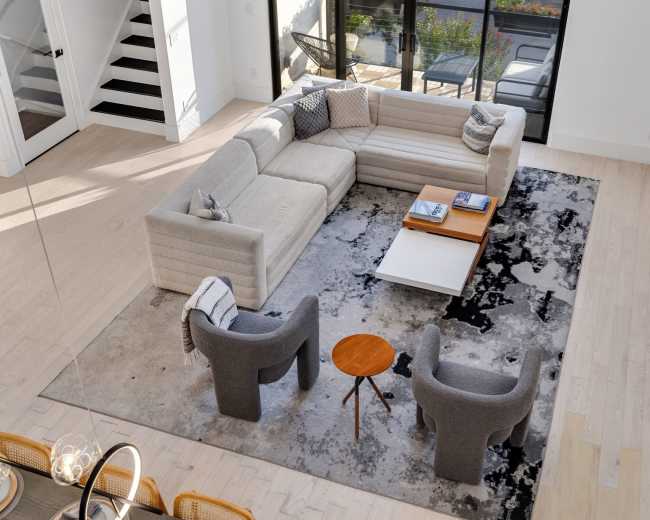 The image shows a modern living room with a large light-colored sectional sofa, two grey armchairs, a small round side table, and a coffee table set on a patterned rug, all arranged around a large window with views of greenery outside.