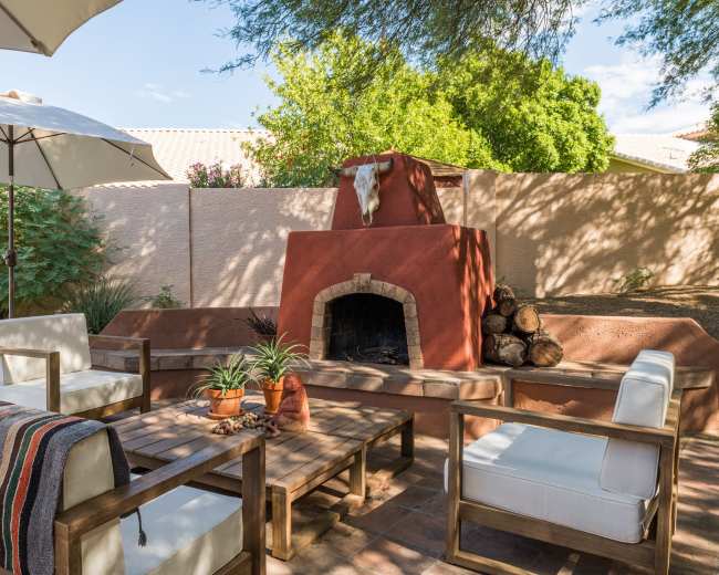The image shows a cozy outdoor seating area with a wooden table and chairs, a fireplace adorned with a decorative bull skull, and an umbrella providing shade.