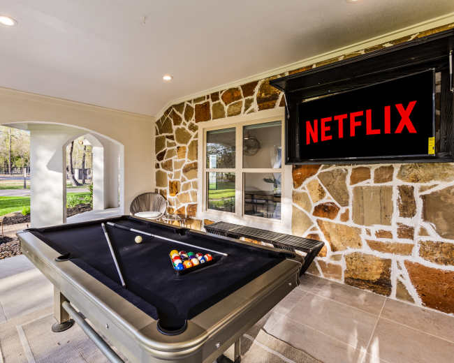 The image shows a game room featuring a billiard table, a large TV displaying the Netflix logo, and a window view of a landscaped outdoor area.