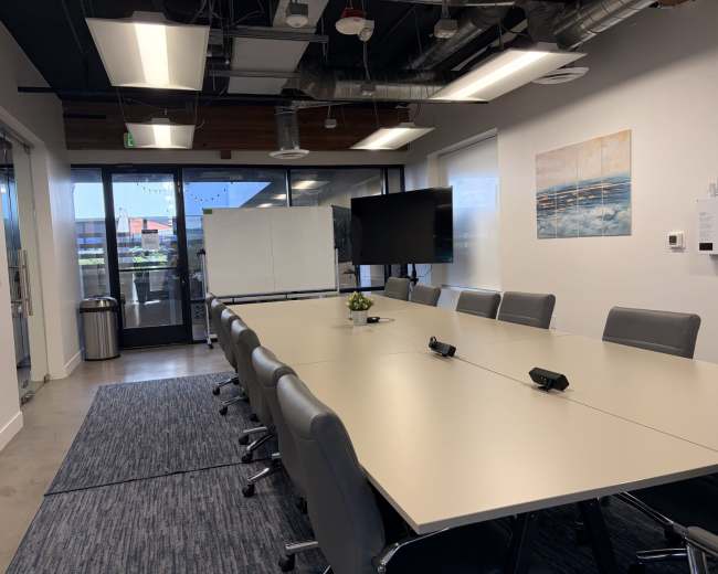 The image shows a modern conference room with a long table, ergonomic chairs, a large screen, and windows that allow natural light.