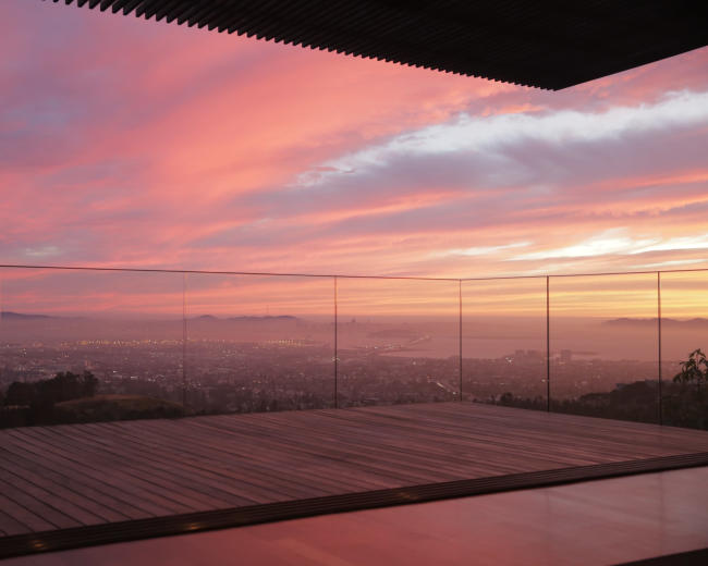 A glass-enclosed balcony overlooking a city skyline at sunset, with vibrant clouds reflecting shades of pink and orange in the sky.