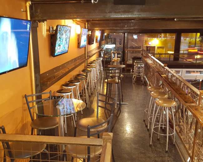 The image shows a narrow bar area with high chairs and tables, featuring multiple television screens mounted on the walls.