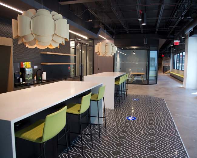 The image shows a modern office break room with a long white countertop, green bar stools, and geometric patterned flooring.