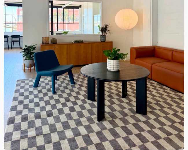 The image shows a modern interior space featuring a brown leather sofa, a round black table, a blue chair, a patterned rug, and large windows letting in natural light.