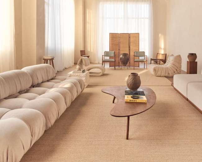 The image shows a minimalist living room with a light-colored interior, featuring a curved sofa, a wooden coffee table, and several pieces of furniture arranged for a spacious feel.