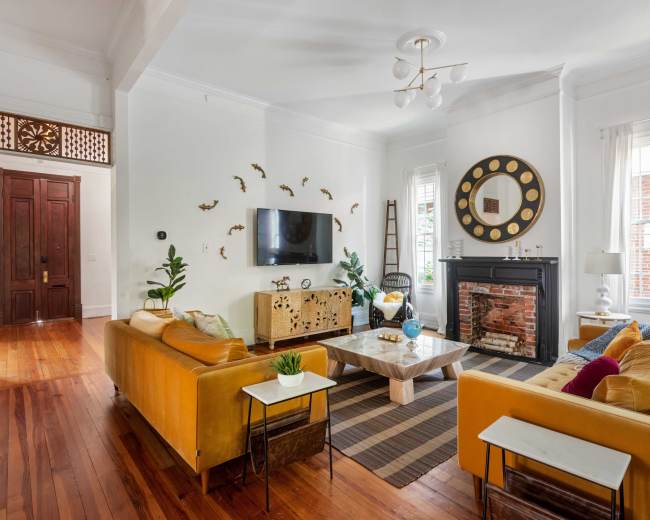 The living room features a combination of yellow sofas, a coffee table, a television mounted on the wall, and a fireplace, with large windows allowing natural light to illuminate the space.