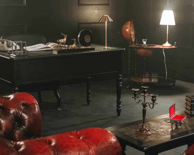 The image depicts a dimly lit office with a wooden desk, leather couches, a globe bar, and a lamp illuminating a side table.