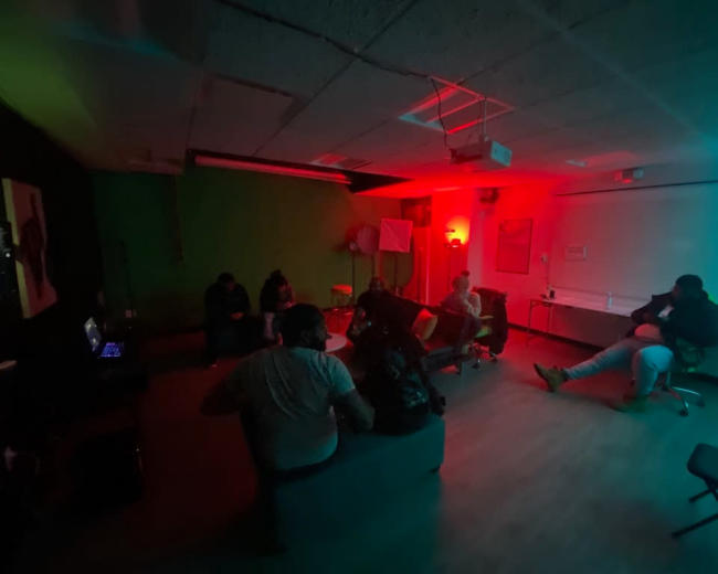 A group of people is gathered in a dimly lit room with red and blue lighting, sitting on various chairs and sofas around a small table.