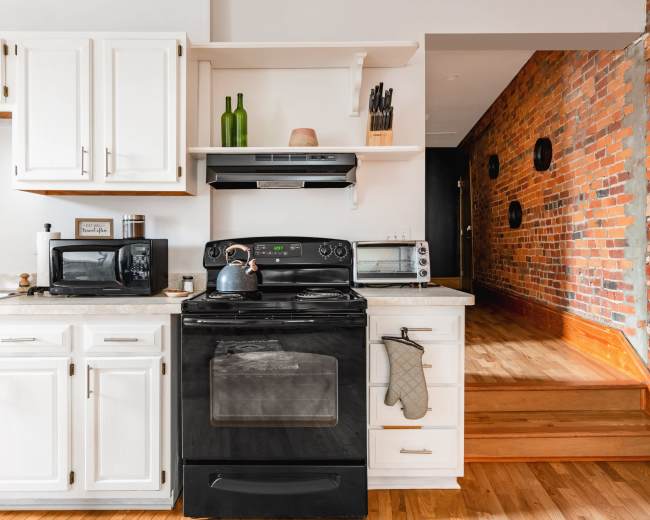 A modern kitchen features white cabinetry, a black stove, a microwave, and a staircase leading to a brick wall area.