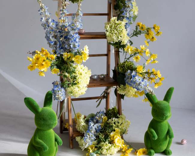 A wooden ladder decorated with colorful flowers is flanked by two green bunny figures and surrounded by decorative eggs.