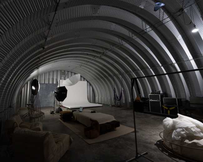 The interior of a curved, industrial space features a photography setup with a backdrop, lighting equipment, and a seating area.