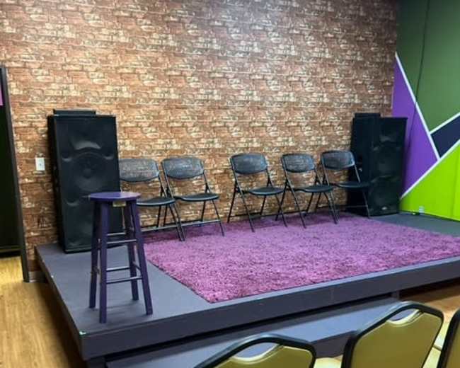 The image shows a small stage with five black folding chairs set up on it, surrounded by a purple rug, and facing an audience area with yellow chairs.