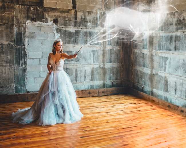 A woman in a flowing dress casts a spell, creating a translucent deer in an empty, rustic room with wooden floors and concrete walls.