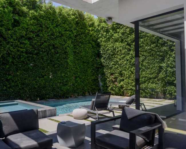A small outdoor patio features black seating, a white side table, and a swimming pool surrounded by tall green hedges.