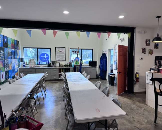 The image shows a spacious art studio with multiple tables set up for painting, colorful bunting hanging from the ceiling, and a counter with supplies along one wall.