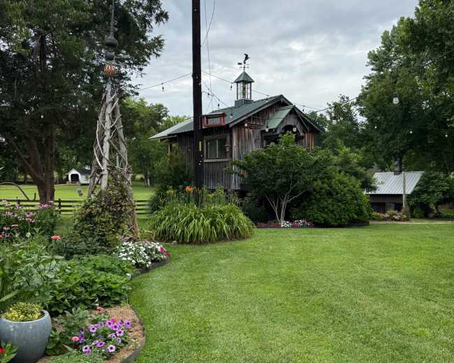 A rustic wooden building with a cupola sits amidst a well-maintained garden featuring flowers and manicured lawns, under a cloudy sky.