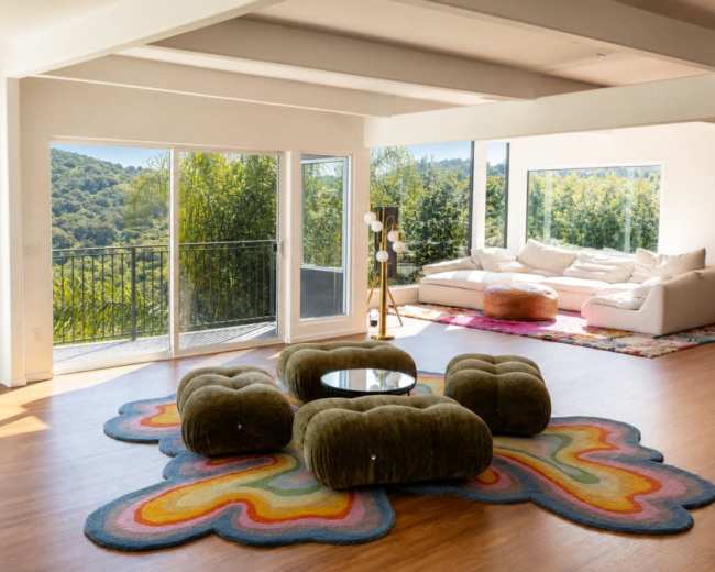 The image shows a spacious living room with large windows overlooking a green hillside, featuring a colorful rug and a white sectional sofa.