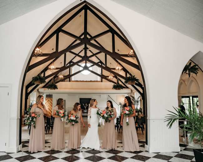 A bride and her bridesmaids stand together in a spacious hall featuring an arched ceiling and patterned tile flooring.