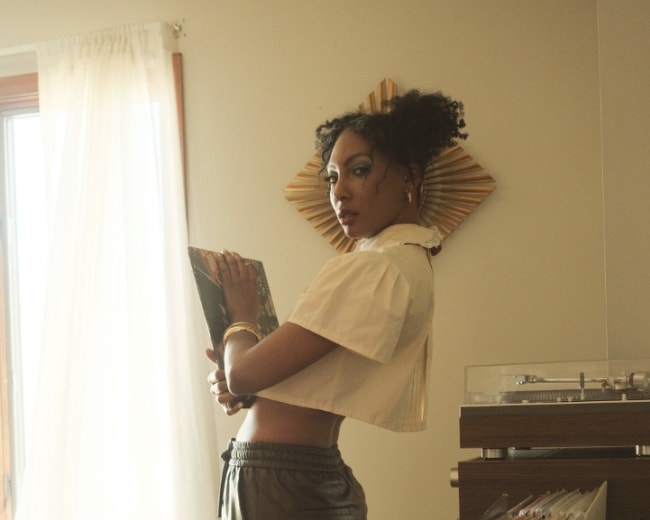 A person with curly hair stands in a room holding a record album, with a row of vinyl records visible in the foreground.