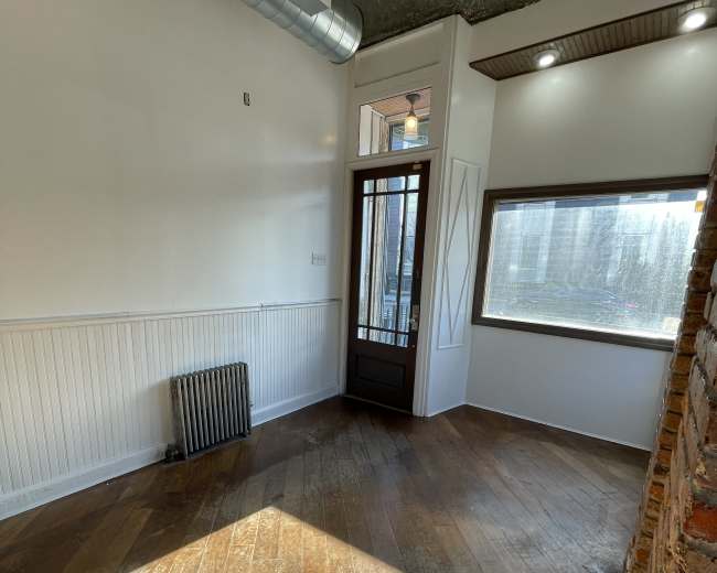 The image shows a small room with wooden flooring, a large window, a door with glass panels, and a radiator against one wall.