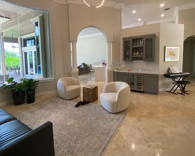 A spacious living area featuring two round armchairs, a small wooden table, and a bar area with cabinetry, all set on a light-colored stone floor.