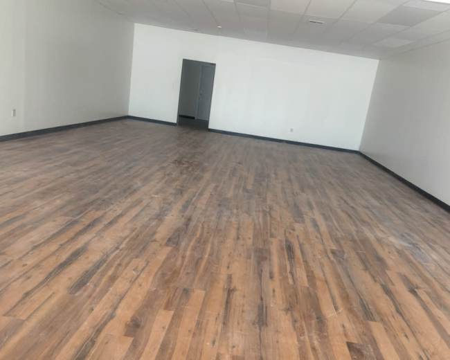 The image shows a spacious, empty room with wooden flooring and plain white walls.