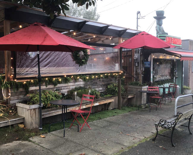 A cozy outdoor seating area featuring red umbrellas and wooden benches, set in front of a restaurant decorated with lights and greenery.