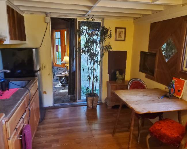 The image shows a small kitchen area with wooden cabinetry, a table and chairs, and a doorway leading to an outdoor view.