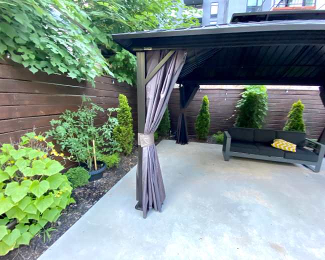 A covered outdoor seating area features a gray sofa and potted plants surrounded by a wooden fence and greenery.