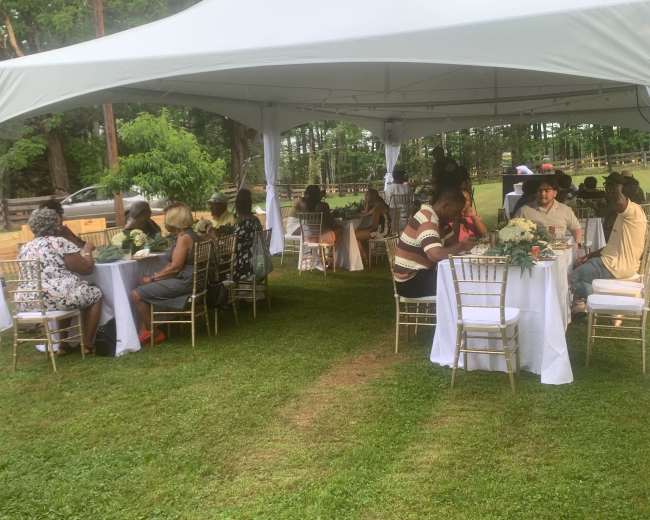 A tented outdoor gathering features several round tables with guests seated, surrounded by greenery.