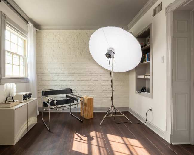 The image shows a well-lit room with a white brick wall, featuring a modern chair, a side table, and a large softbox light on a stand.