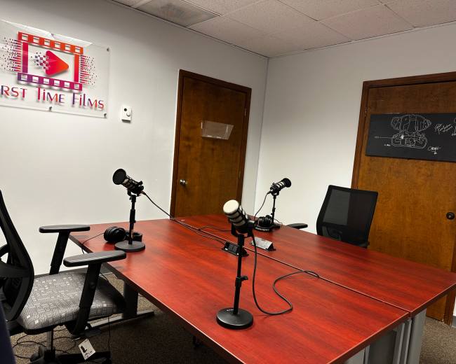 The image shows a small recording studio with a red table, three microphones, and a blackboard on the wall, alongside wooden doors and a sign that reads "First Time Films."