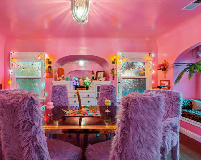 A pink-themed dining area features a table surrounded by fluffy chairs, illuminated by decorative lights and a central pendant fixture.