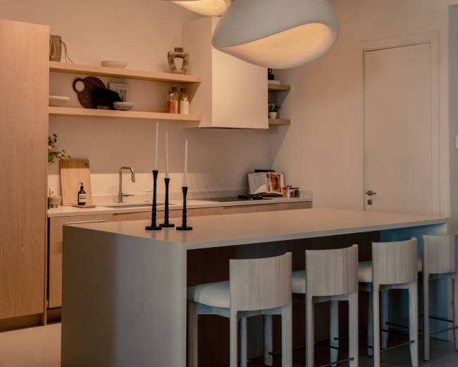 The image shows a modern kitchen with a large island, minimalist cabinetry, and pendant lighting, featuring bar stools along one side.