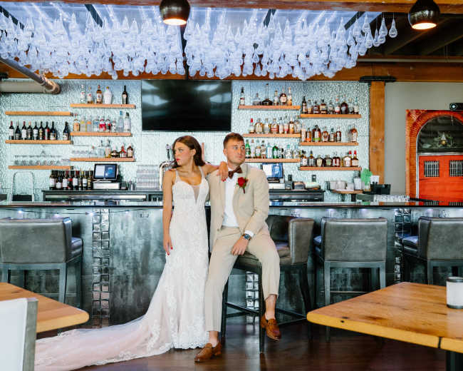 A bride in a lace wedding dress stands beside a groom in a beige suit at a bar with an array of bottles displayed on shelves behind them.