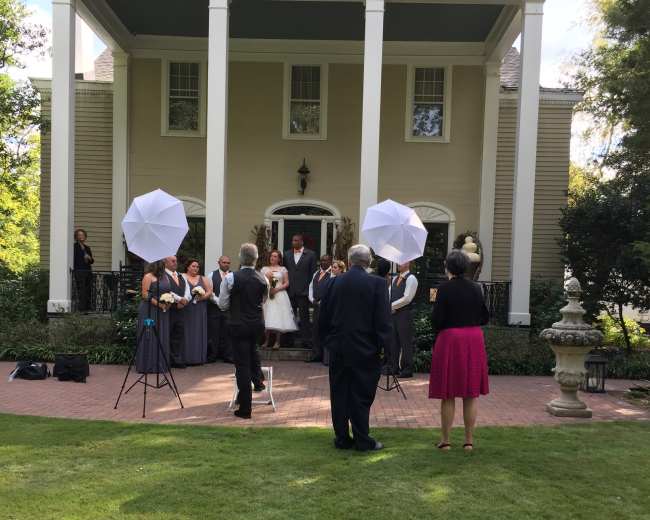 A group of people is gathered outside a large house for a formal photo shoot, with several individuals holding umbrellas for lighting and others standing in a semi-circle.