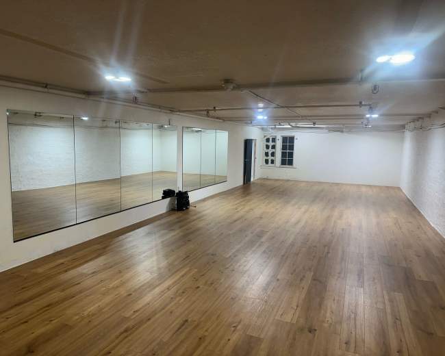 The image shows an empty dance studio with wooden flooring and large mirrors on one wall.