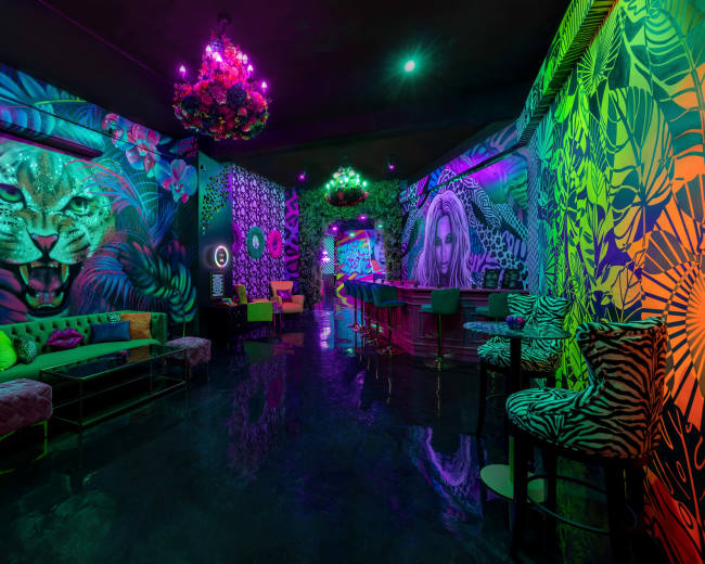 The image features a vibrant, neon-lit lounge with colorful murals of a leopard and a woman, adorned with plush seating and a bar.