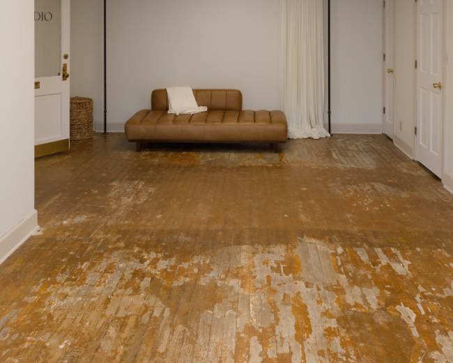 The image shows a spacious room with a brown sofa, wooden floor showing signs of wear, and white walls with a draped curtain and a light fixture on the ceiling.