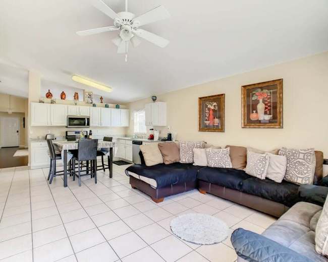 The image shows a spacious, open-concept living area with a sectional sofa, a dining table, and a kitchen with white cabinets.