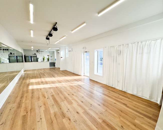 The image shows a spacious room with wooden flooring, white walls, and large windows, featuring a mirror along one side and sheer curtains.